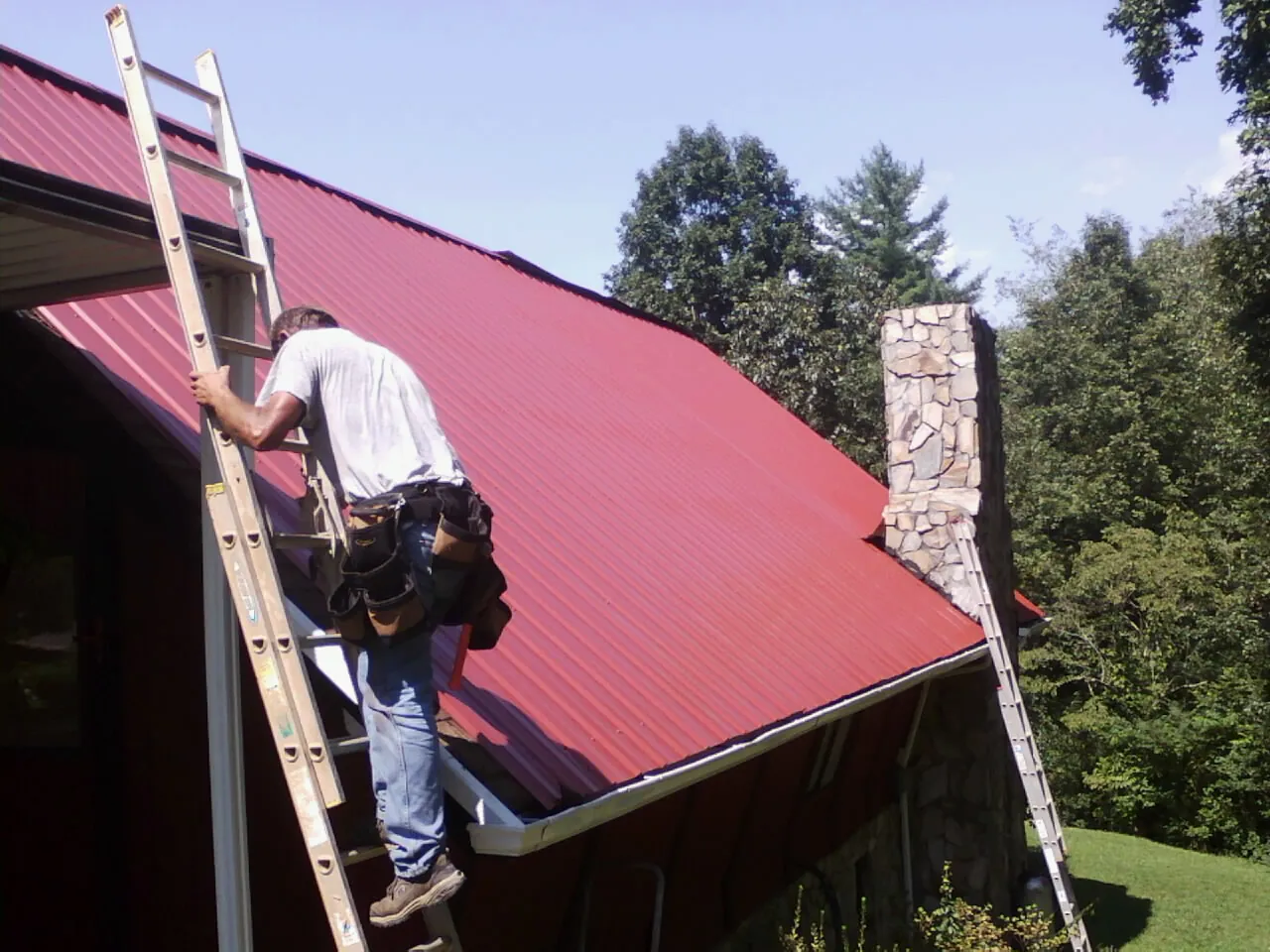 Roofer on a ladder beside a red metal roof and stone chimney — Soffit & Fascia Repair by Roofing Brothers in Bobtown